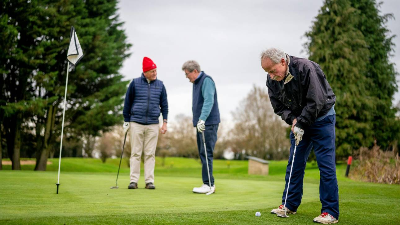 Three Genetlman playing golf. One in foreground putting, two in background talking
